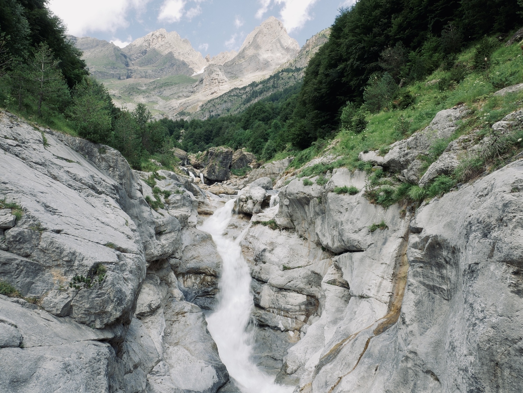 A waterfall cascades through rocky cliffs surrounded by lush green vegetation and mountains in the background.